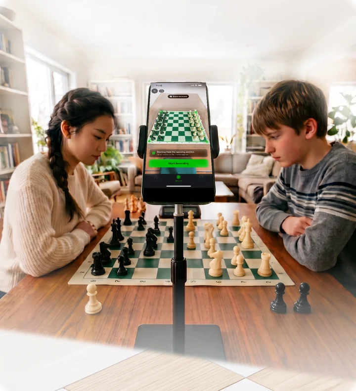 Two players at a board with a phone on a stand between them, framing the position.